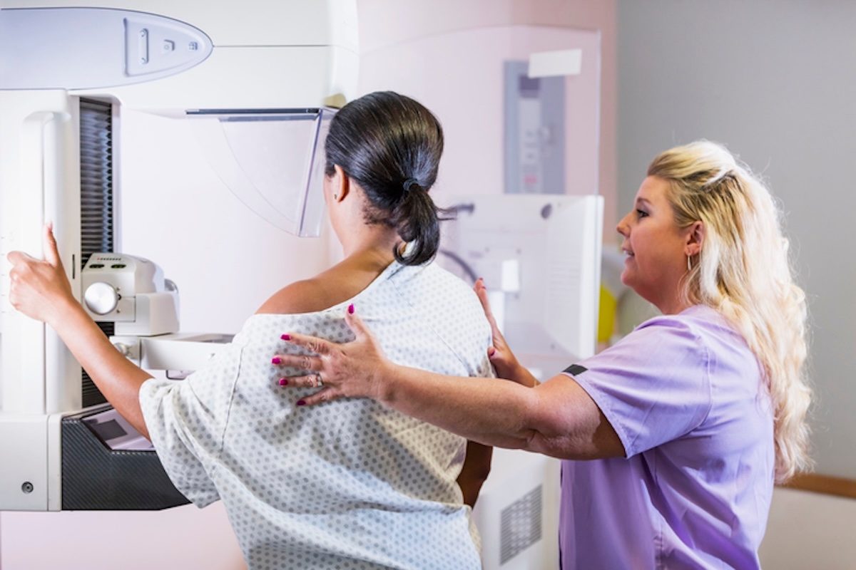 dark haired woman getting a mammogram with her back toward us, light haired woman assisting her