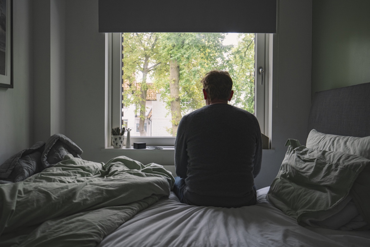 Man sitting on a bed looking out a window, appearing thoughtful or sad.