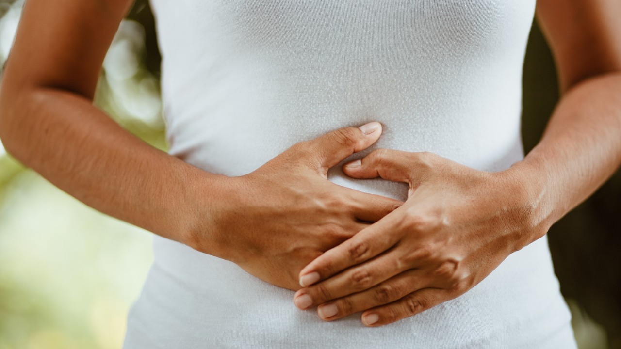 Person in a white shirt holding their hands over their stomach, standing outdoors with blurred greenery in the background.