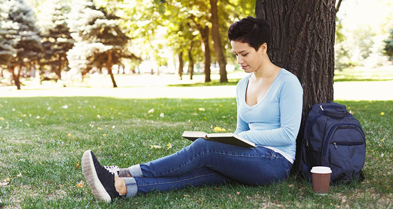 Woman sitting against a tree in a park, reading a book with a backpack and coffee cup beside her.