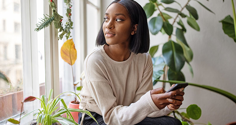 Woman holding a smartphone while sitting by a window, surrounded by green plants, gazing outside with a thoughtful expression.