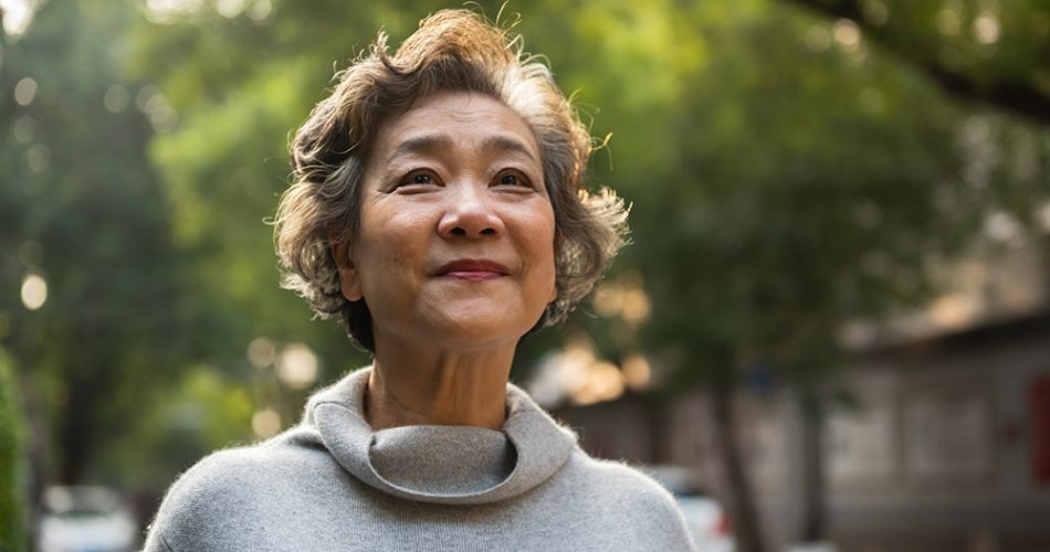 Older woman with short, wavy gray hair standing outdoors, looking into the distance with a peaceful expression, surrounded by green trees.