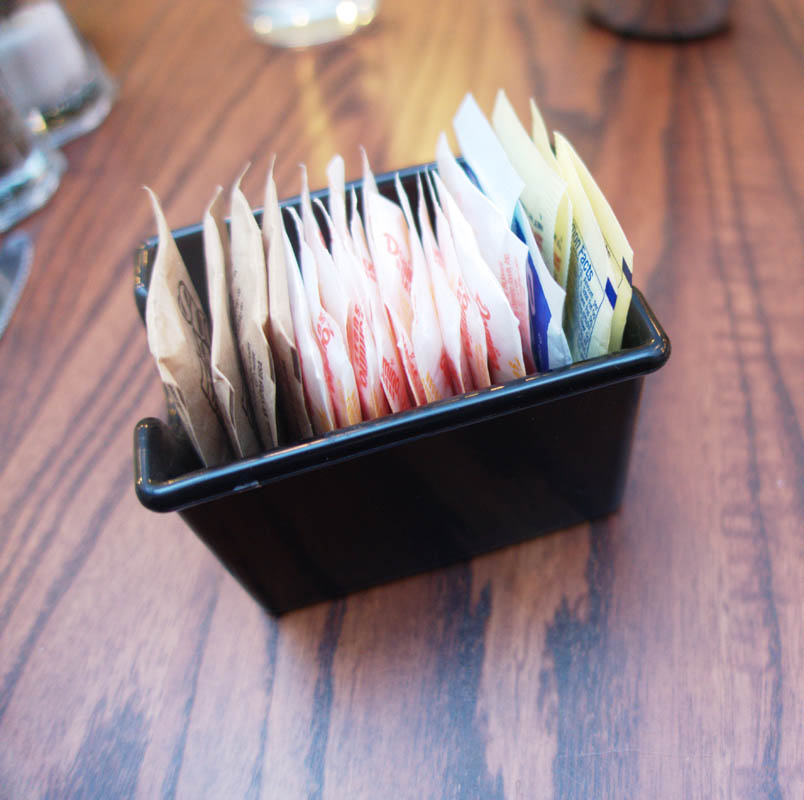 Small black container filled with various sugar and sweetener packets, neatly arranged on a wooden table.