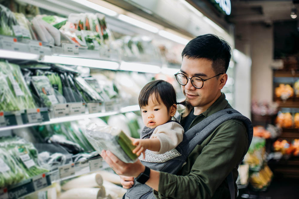 Father holding his young child in a baby carrier while grocery shopping, both looking at a package of fresh vegetables in the produce section.