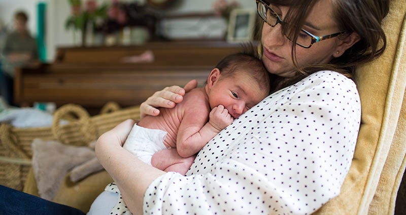 Mother in glasses and a polka dot blouse cradling her newborn baby on her chest in a cozy home setting, with a piano and wicker furniture in the background.