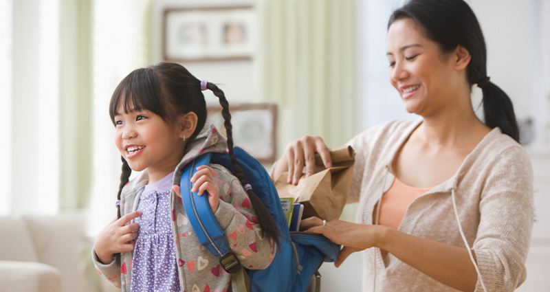 A smiling mother packs a lunch into her young daughter’s blue backpack as the girl, wearing a hoodie with braids, excitedly prepares for school. The background features a bright and cozy living room.