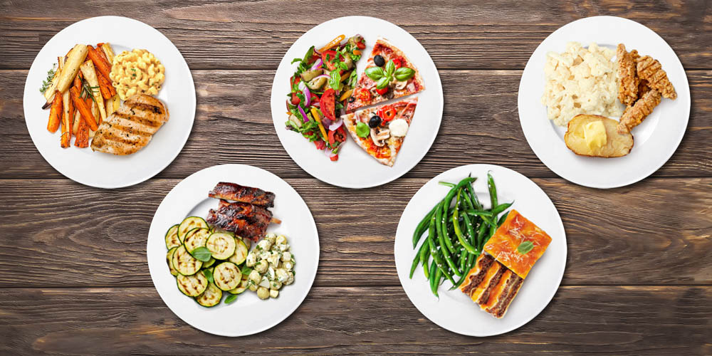 Five plates of food on a wooden table: grilled chicken, veggie pizza, steak with veggies, green beans with fish, and fried chicken with mashed potatoes.