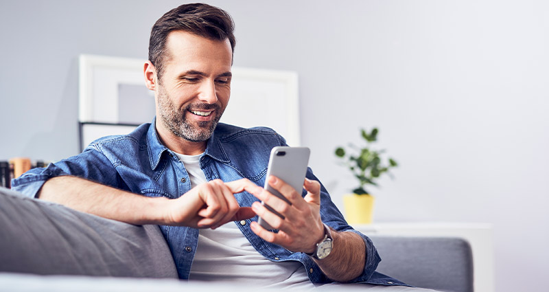  A man sitting on a couch, smiling while using a smartphone, with a modern living room in the background.
