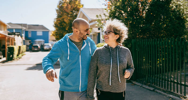  Two smiling people walking together on a sunny street, one in a light blue hoodie and the other in a gray jacket.