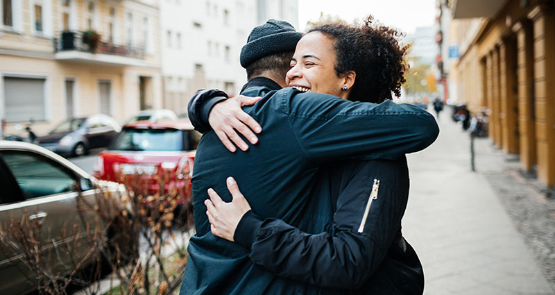 Two people embracing on a city street, smiling and enjoying the moment. One person is wearing a black jacket and the other is in a dark blue jacket.