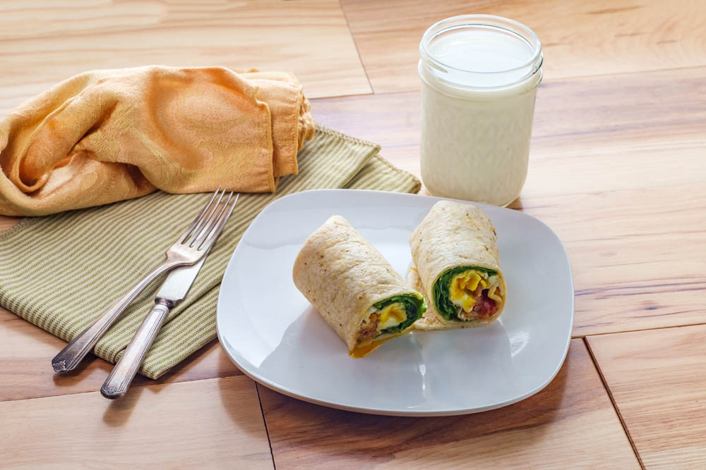 A breakfast wrap cut in half on a white plate, next to a glass of milk, with a fork, knife, and cloth napkin on a wooden surface.
