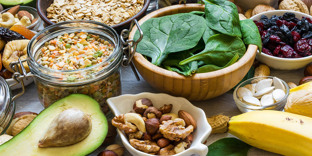 A variety of healthy foods displayed on a table, including nuts, fruits, spinach, and grains, arranged in bowls and containers.