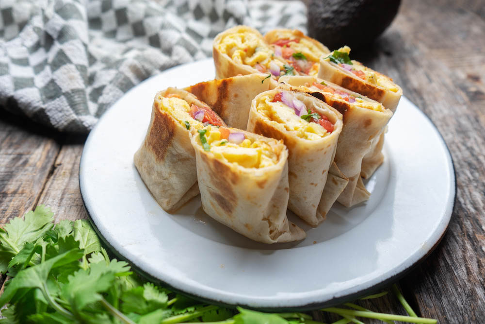 A plate of sliced breakfast burritos filled with eggs, tomatoes, and herbs, arranged neatly, with fresh cilantro and a checkered napkin in the background on a rustic wooden surface.