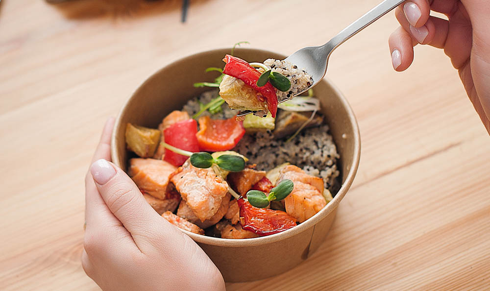 A person's hand holding a fork above a bowl filled with salmon, vegetables, and rice, placed on a wooden table.