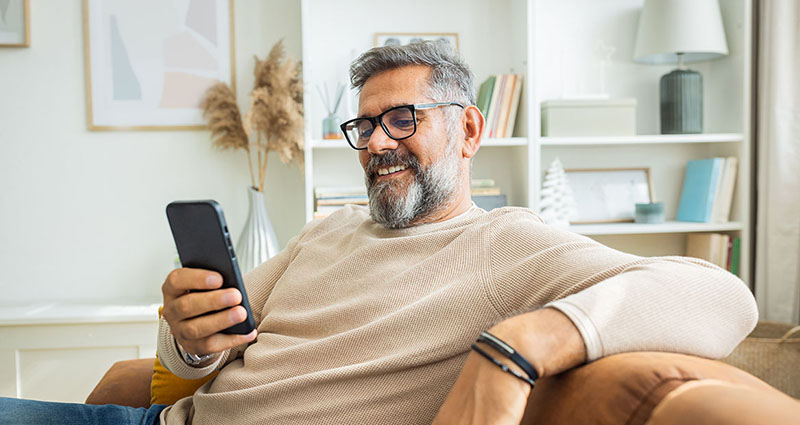  A smiling older man with glasses sitting on a couch, looking at his smartphone in a cozy living room setting.