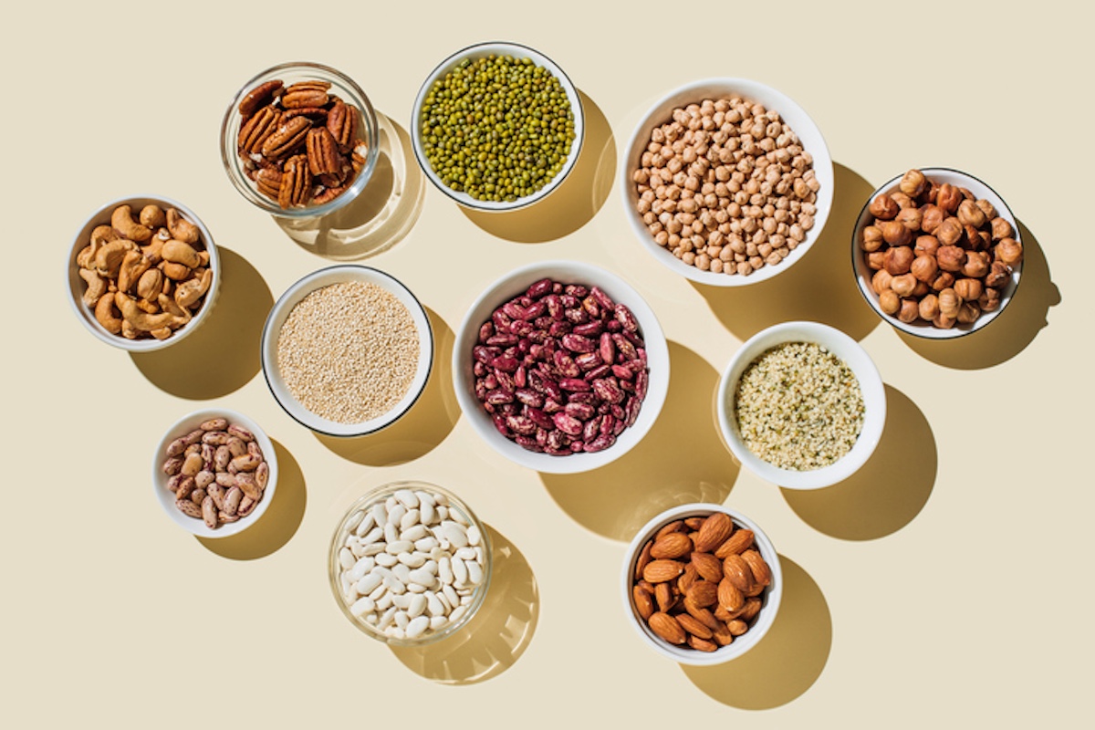 Overhead view of eleven small bowls of different nuts, legumes, and grains on a nuetral background.