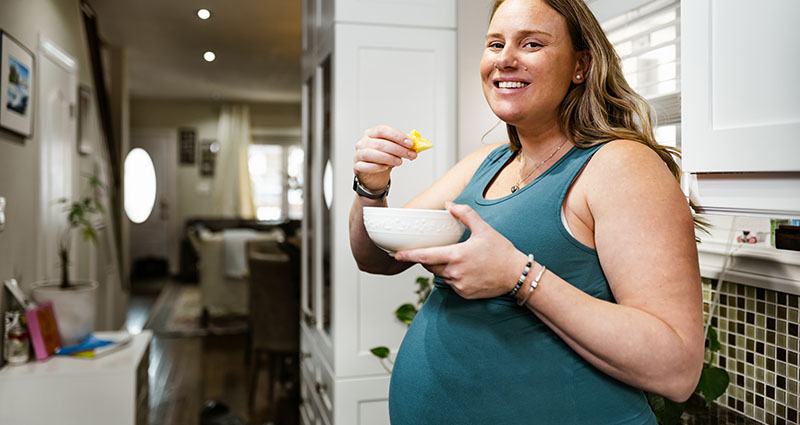 A pregnant woman in a tank top smiling while holding a bowl and eating, standing in a warmly lit kitchen.