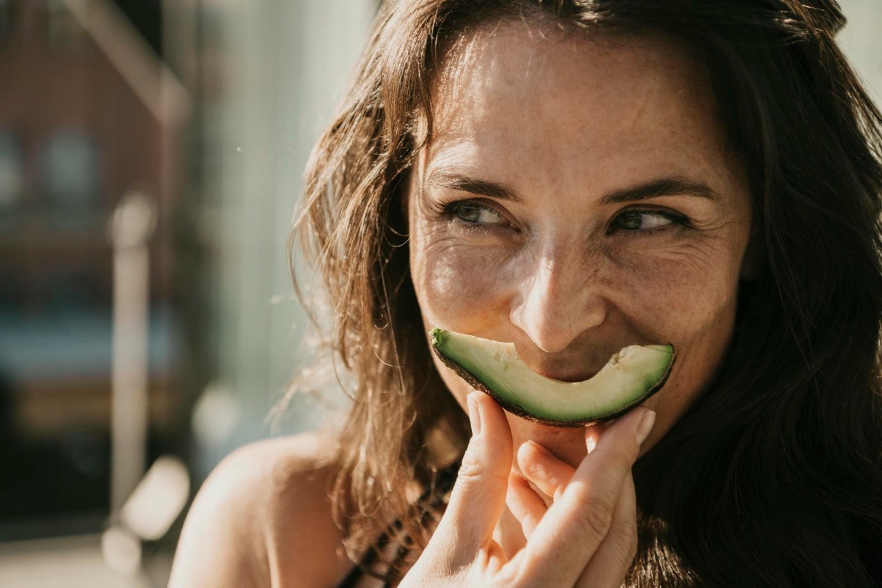A woman smiling playfully while holding a slice of cucumber in front of her mouth, with a bright outdoor background.