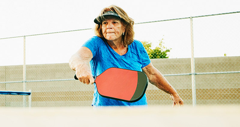 An older woman wearing a blue shirt and a visor is playing pickleball, holding a paddle and preparing to hit the ball, with a fence and trees in the background.