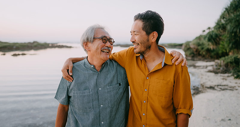 Two men, one older and one younger, are smiling and embracing each other by a body of water during sunset, with a natural landscape in the background.