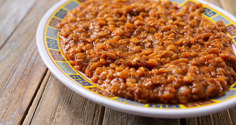 A plate of cooked lentils in a thick, brown sauce on a decorative plate, placed on a wooden surface.