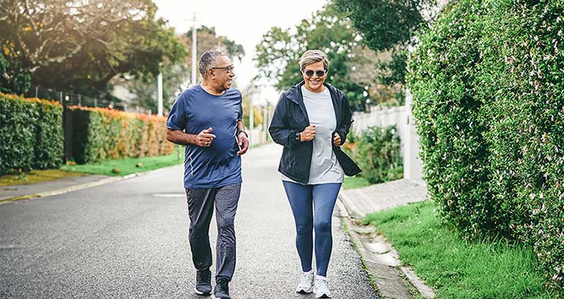 Two older adults walking together on a tree-lined street, smiling and chatting, dressed in casual athletic wear.