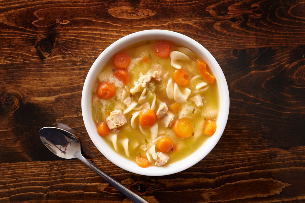 A bowl of chicken noodle soup with carrots, noodles, and chicken pieces on a wooden table, next to a spoon.