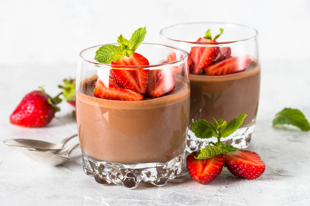 Two glasses of chocolate mousse topped with sliced strawberries and mint leaves, with extra strawberries and a spoon on the side.