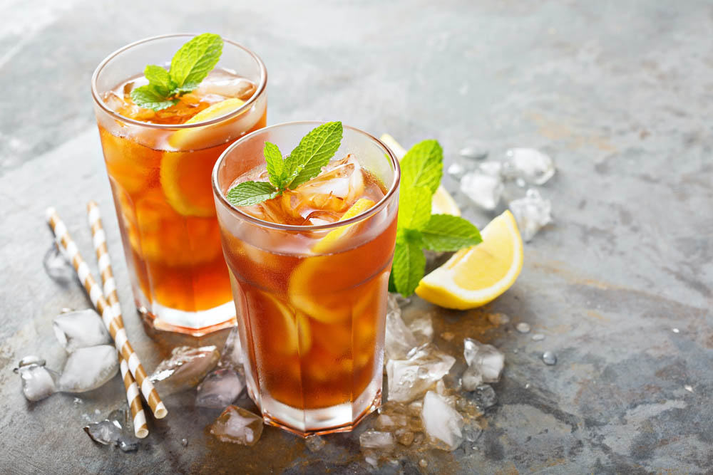 Two glasses of iced tea with lemon slices and mint leaves, surrounded by ice cubes and lemon wedges on a textured surface.
