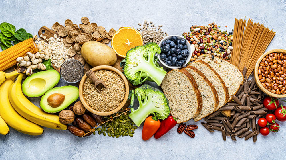 Overhead view of a large group of food with high content of dietary fiber arranged side by side. The composition includes berries, oranges, avocado, chia seeds, wholegrain bread, wholegrain pasta, whole wheat, potatoes, oat, corn, mixed beans, brazil nut, sunflower seeds, pumpkin seeds, broccoli, pistachio, banana among others. 