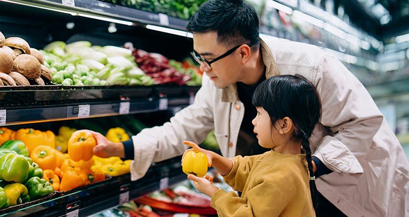 An adult and a child are shopping for vegetables in a grocery store, with the adult reaching for a yellow bell pepper while the child watches.