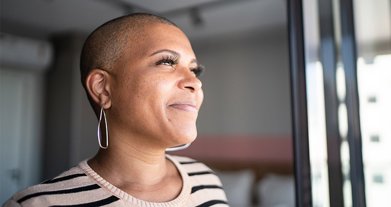 A woman with a shaved head and large hoop earrings is smiling and looking out of a window, with soft natural light illuminating her face.