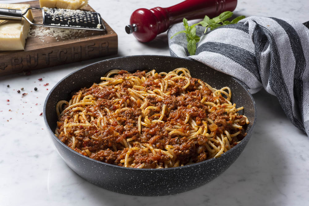 A bowl of spaghetti with meat sauce on a marble counter, garnished with grated cheese and a red pepper mill nearby.