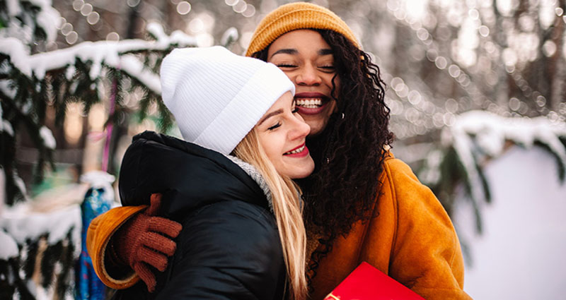 Two women are embracing and smiling in a snowy outdoor setting, wearing winter clothing, with trees covered in snow in the background.
