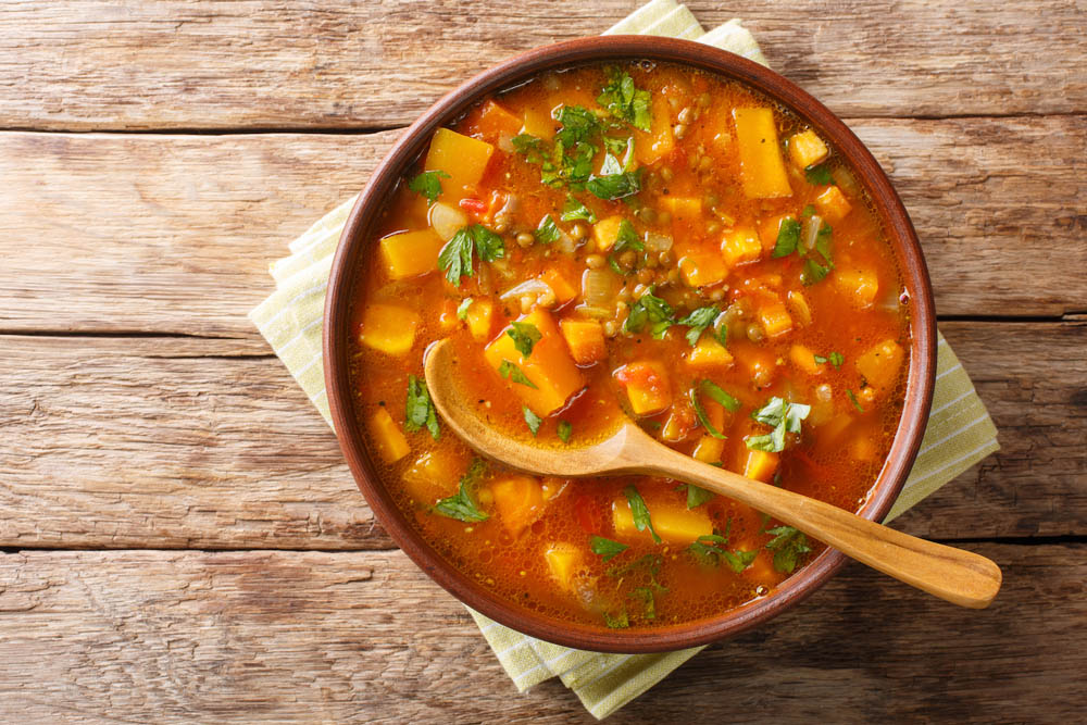 A bowl of vegetable soup with lentils, garnished with parsley, and a wooden spoon on a wooden table.