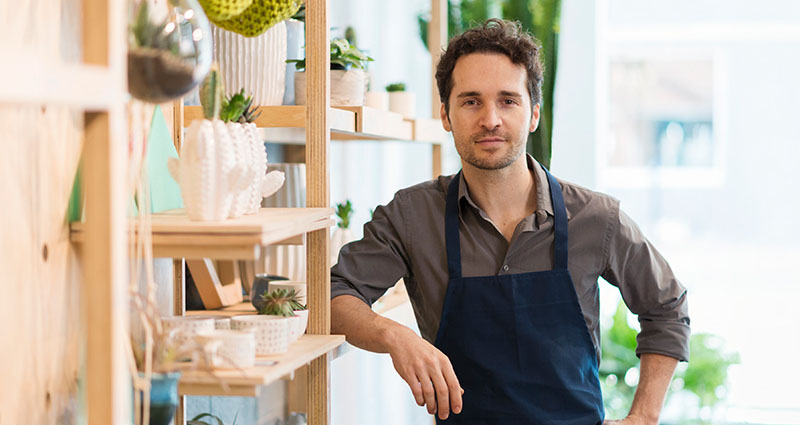 A young man in an apron is standing in a bright, modern shop filled with plants and home goods, smiling at the camera.