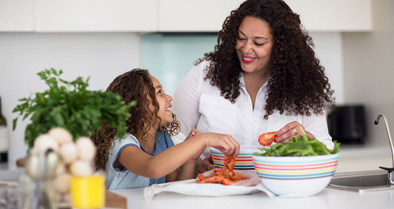A woman and a young girl are preparing food together in a bright kitchen, with fresh ingredients and a large bowl in front of them, both smiling and engaged in the activity.