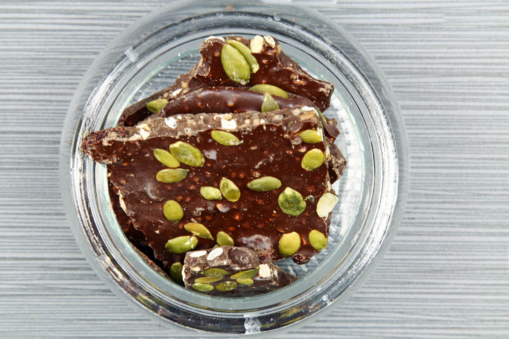 Glass jar containing pieces of chocolate bark topped with pumpkin seeds, viewed from above on a striped surface.