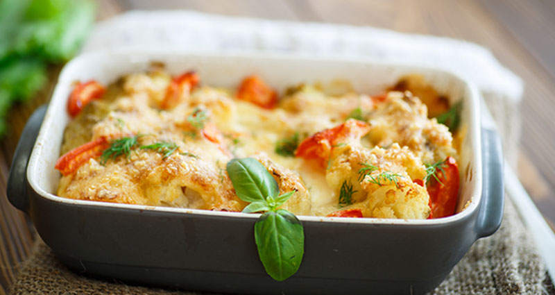 A close-up of a baked dish in a rectangular pan, topped with melted cheese, vegetables, and garnished with fresh basil leaves.
