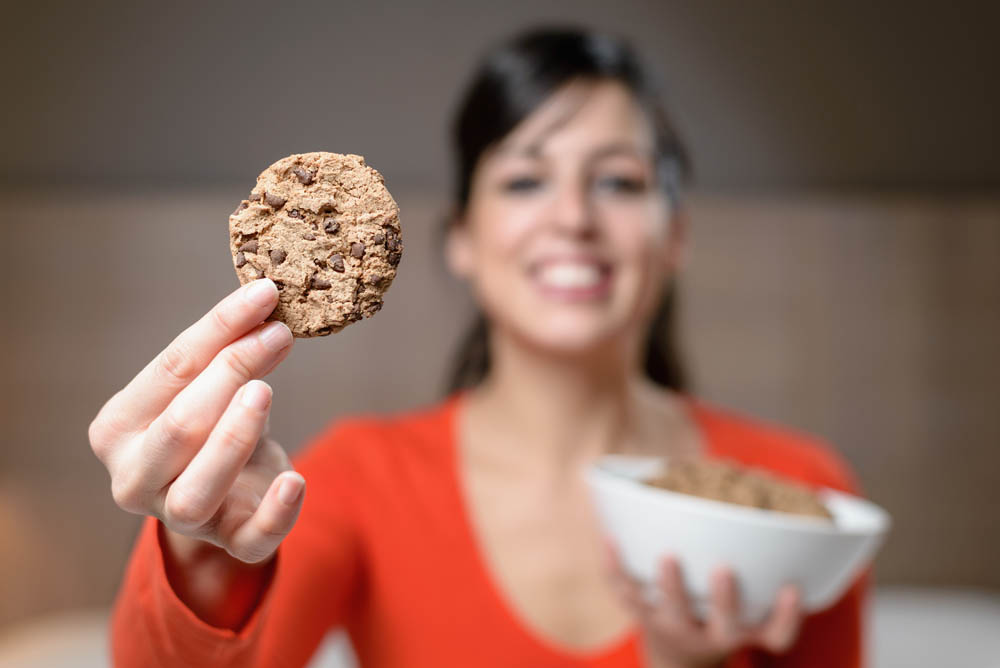 Woman showing a cookie and holding a bowl. Night scene with hungry cheerful brunette caucasian girl in her room.