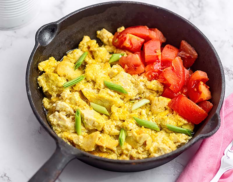 Scrambled eggs with green onions and diced tomatoes in a black skillet on a marble surface.
