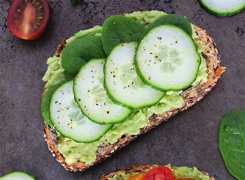 A slice of multigrain bread topped with mashed avocado, fresh cucumber slices, and spinach leaves, on a dark surface.
