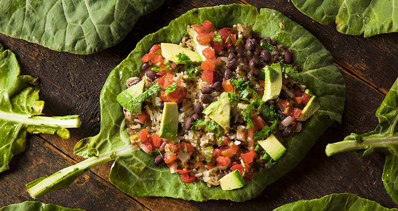 Leaf wrap with diced tomatoes, black beans, rice, avocado, and herbs on a wooden surface, surrounded by other leaves.