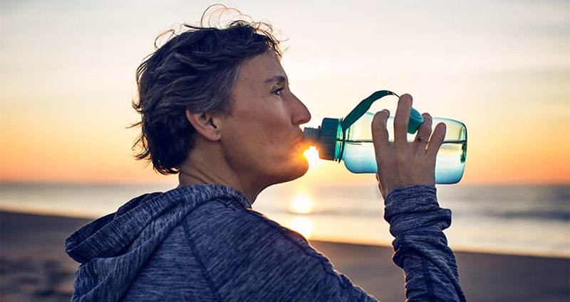 Woman drinking water from a bottle at the beach during sunset.