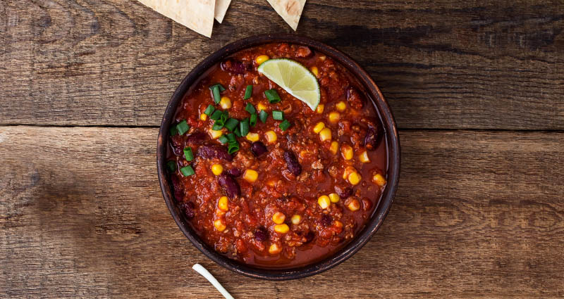 A bowl of chili with corn, beans, and a lime wedge, garnished with green onions, on a wooden surface. Crackers are nearby.