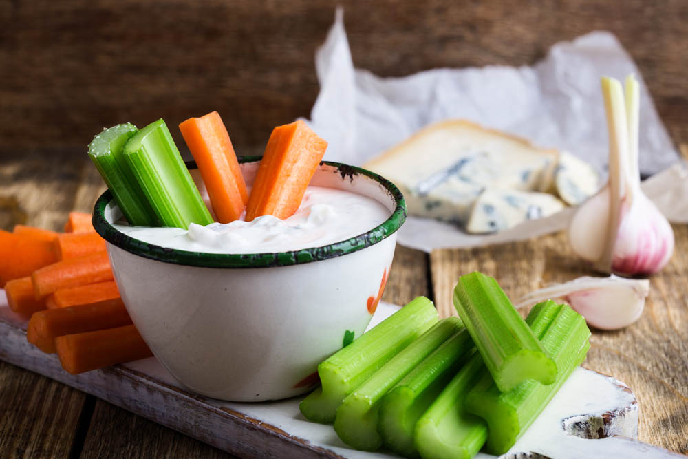 Bowl of creamy dip with celery and carrot sticks on a wooden board, with blue cheese and garlic in the background.