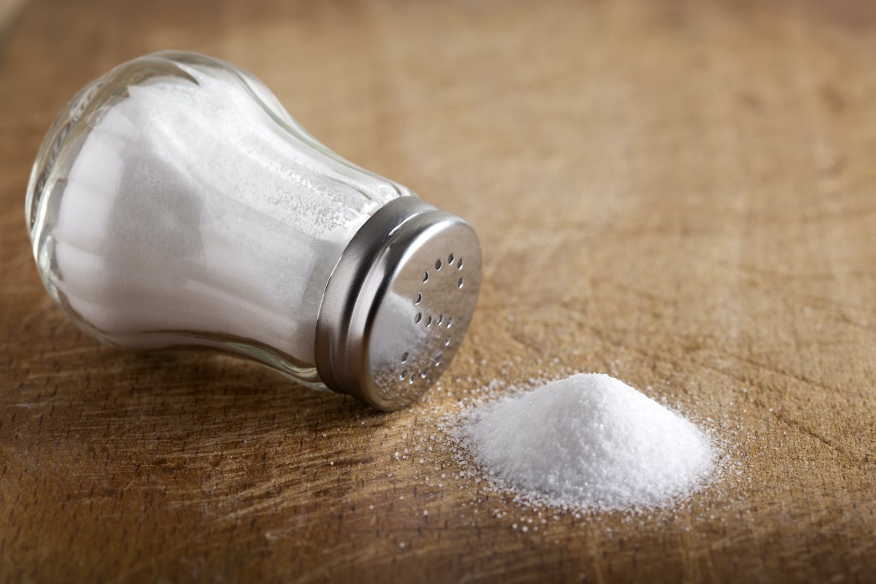A tilted salt shaker lies on its side on a wooden surface, with a small mound of spilled salt next to it.