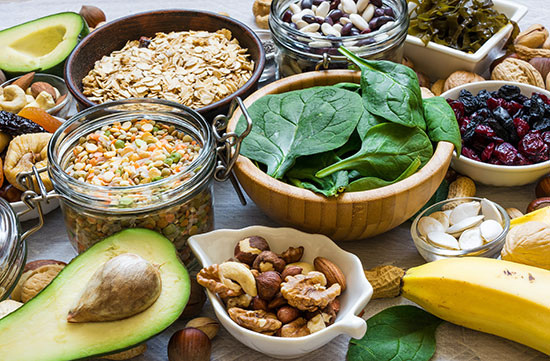 Variety of healthy foods including spinach, nuts, seeds, banana, avocado, oats, and dried fruits arranged on a table.