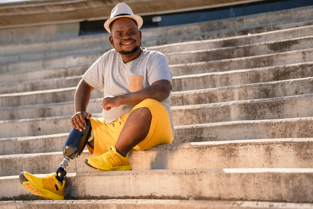 Man with a prosthetic leg sits on concrete steps, wearing a hat, gray T-shirt, yellow shorts, and sneakers, smiling at the camera.