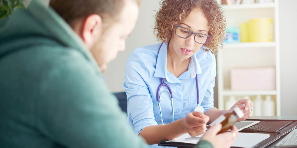  A healthcare professional with curly hair and glasses discusses information with a patient at a table, holding a smartphone.
